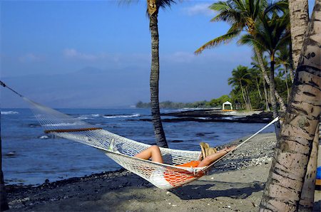 Vacationer relaxes in a hammock on the Kohala Coast.  View includes Mauna Kea mountains and the Makaiwa Bay Beach.  She is wearing a straw hat, orange shirt, white shorts and tennis shoes. Foto de stock - Royalty-Free Super Valor e Assinatura, Número: 400-07829416