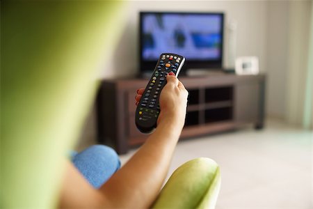 Over the shoulder view of girl sitting on sofa holding tv remote and surfing programs on television Foto de stock - Super Valor sin royalties y Suscripción, Código: 400-07903243