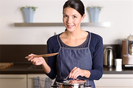 simsearch:400-08021946,k - Young woman cooking the food for dinner over the stove in her kitchen standing holding the lid of a stainless steel saucepan and wooden ladle as she smiles at the camera Stock Photo - Budget Royalty-Free & Subscription, Code: 400-07992947