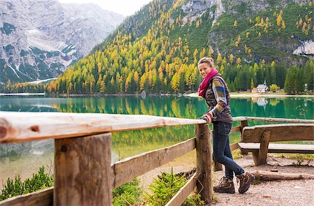 A smiling brunette hiker wearing outdoor gear stands facing the water and leaning against a wooden railing. The still water in the background provides a perfect mirror image of the mountains and trees. The autumn colours are golden. Foto de stock - Super Valor sin royalties y Suscripción, Código: 400-08054027
