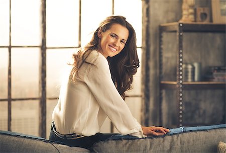 simsearch:400-08054469,k - An elegant brunette sitting on the back of sofa in a loft, leaning forward and smiling is looking over her shoulder. In the background, the daylight is streaming through a window. Chic loft. Stock Photo - Budget Royalty-Free & Subscription, Code: 400-08054456