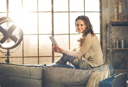 simsearch:400-08054488,k - A brunette woman is smiling, looking up from her  tablet PC while sitting on the back of a sofa. Industrial chic ambiance and cozy atmosphere, sunlight is streaming through the loft window. Stock Photo - Budget Royalty-Free & Subscription, Code: 400-08054468