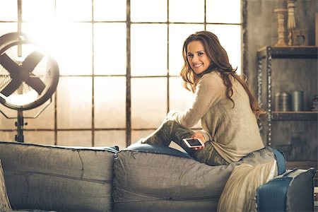 simsearch:400-08054469,k - A brunette woman is smiling with phone sitting on the back of a sofa. Industrial chic ambiance and cozy atmosphere, sunlight is streaming through the loft window. Stock Photo - Budget Royalty-Free & Subscription, Code: 400-08054469