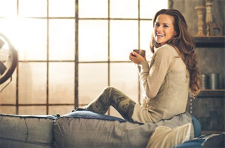 simsearch:400-08054469,k - A brunette long-haired woman is seen from the side while sitting on the back of a sofa. She  is smiling and holding a hot cup of coffee. Industrial chic background, and cozy atmosphere. Stock Photo - Budget Royalty-Free & Subscription, Code: 400-08054478