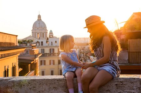 At sunset, a brunette mother wearing summer clothing and a hat and a blonde daughter are sitting holding hands and talking while sitting on a ledge. In the distance, St. Peter's Basilica. Stock Photo - Budget Royalty-Free & Subscription, Code: 400-08134098