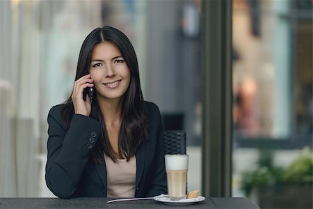 Smiling woman chatting on her mobile phone while sitting in a cafeteria enjoying a delicious cup of cappuccino coffee giving the camera a friendly smile Stock Photo - Budget Royalty-Free & Subscription, Code: 400-08160558