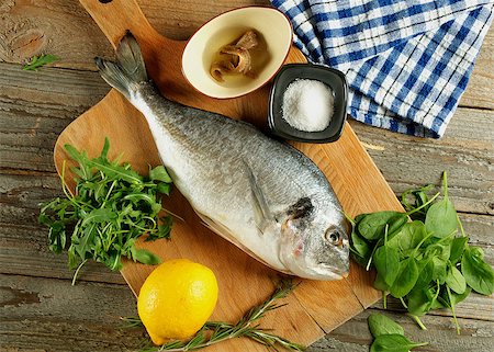 Cooking Raw Dorado Fish with Arugula, Herbs, Spices, Anchovies and Lemon on Wooden Cutting Board closeup. Top View Foto de stock - Super Valor sin royalties y Suscripción, Código: 400-08161951