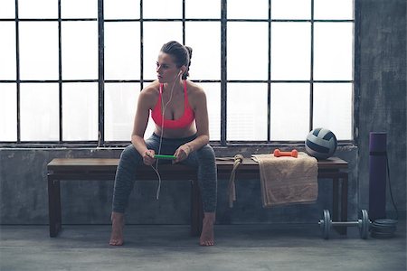 simsearch:400-08189778,k - Looking off to the side, an athletic woman watches something in the distance. She is sitting on a bench by a window in a loft gym, holding her device and listening to music. Foto de stock - Super Valor sin royalties y Suscripción, Código: 400-08189772