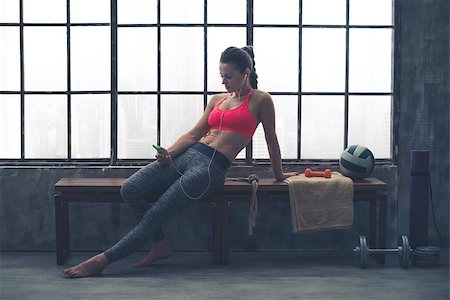 simsearch:400-08189778,k - A fit, buff, muscular woman is sitting, relaxed, on a wooden bench in her local loft gym. Looking down at her phone, she is selecting the music she would like to listen to for her workout. Foto de stock - Super Valor sin royalties y Suscripción, Código: 400-08189780