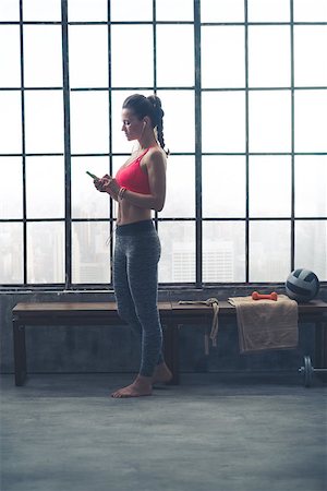 simsearch:400-08189778,k - Standing by a wooden bench which is placed by a big window, a fit, healthy, strong young woman is looking down at her device and selecting which music to listen to for her workout. Foto de stock - Super Valor sin royalties y Suscripción, Código: 400-08189781