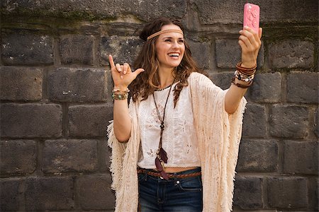 simsearch:400-08194027,k - Longhaired hippy-looking young lady in jeans shorts, knitted shawl and white blouse standing near stone wall in old town and making selfie Photographie de stock - Aubaine LD & Abonnement, Code: 400-08195206