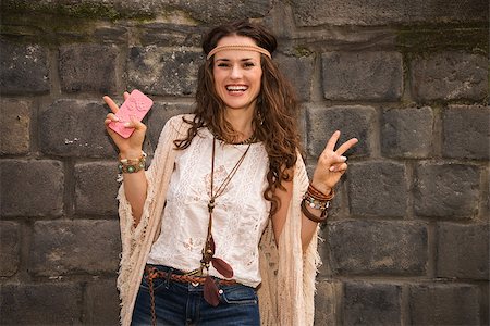 simsearch:400-08194027,k - Longhaired hippy-looking young lady in jeans shorts, knitted shawl and white blouse standing near stone wall and showing victory gesture Photographie de stock - Aubaine LD & Abonnement, Code: 400-08195210