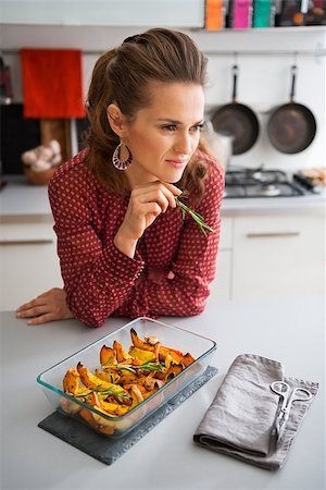 simsearch:400-07956018,k - An elegant woman cook leans against the kitchen counter, thinking about the next ingredients. In her hand, she holds a sprig of rosemary. On the counter, hot roasted pumpkin is cooling. Stockbilder - Microstock & Abonnement, Bildnummer: 400-08196817
