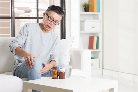 simsearch:400-08318516,k - Portrait of casual 50s mature Asian man knee pain, pressing on knee joint with painful expression, sitting on sofa at home, medicines and water on table. Stockbilder - Microstock & Abonnement, Bildnummer: 400-08225189