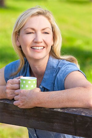 simsearch:400-08299085,k - Beautiful happy middle aged woman in her thirties or forties smiling and leaning on fence in sunshine drinking tea or coffee Stock Photo - Budget Royalty-Free & Subscription, Code: 400-08294702