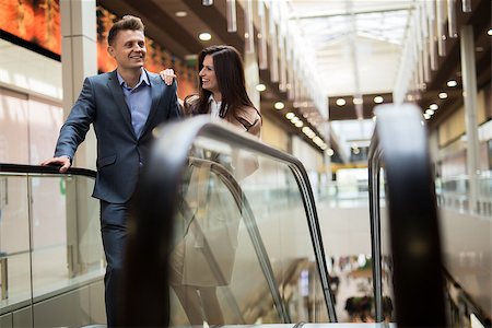 Beautiful young couple on the escalator at the mall. Dark-haired girl in a beige coat Beautiful smiling. Blonde man in a luxury suit smiling at her. Arrived shopping Stock Photo - Budget Royalty-Free & Subscription, Code: 400-08345269