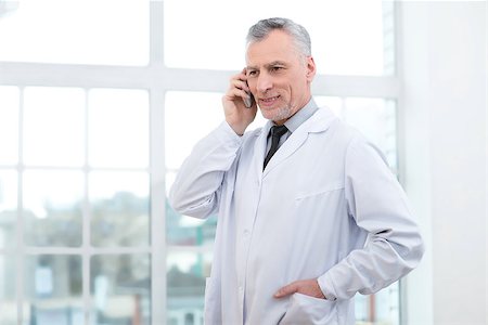 Portrait of aged doctor wearing lab coat. Doctor in years standing in hospital office with big window. Medico smiling and using mobile phone Stock Photo - Budget Royalty-Free & Subscription, Code: 400-08376733