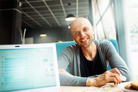 joyful smiling man sitting in a cafe near the open laptop at a table made of wood and holds a smart phone with headphones. in the background a bright window with bright daylight Fotografie stock - Microstock e Abbonamento, Codice: 400-08671225