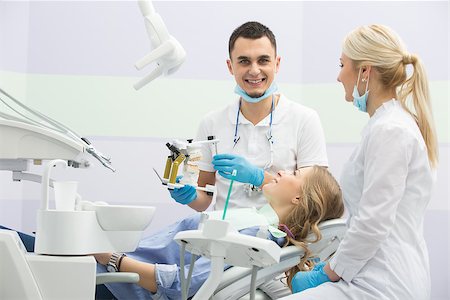 dentist bib girl - Pretty smiling girl in blue shirt and patient bib on the patient chair in the dental cabinet. Near her sits a smiling male dentist and a female assistant. They both wear white uniform with blue latex gloves and blue masks. Dentist also has binocular loupes. He holds an articulator with teeth mould. Horizontal. Stock Photo - Budget Royalty-Free & Subscription, Code: 400-08706061