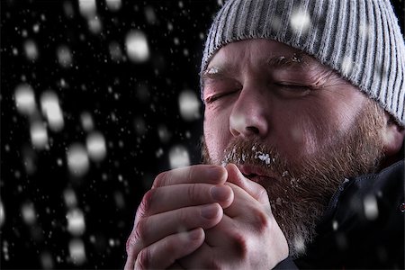 Freezing cold man standing in a snow storm blizzard trying to keep warm. Eyes closed and blowing warm air into his hands. Wearing a beanie hat and winter coat with frost and ice on his beard and eyebrows. Looking to the left. Foto de stock - Royalty-Free Super Valor e Assinatura, Número: 400-08795864