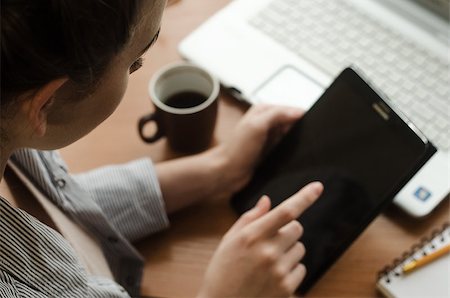 Young woman sitting at a laptop and holding a a tablet in hands. Nearby stands a a cup of coffee and lie notebook with pencil Foto de stock - Super Valor sin royalties y Suscripción, Código: 400-08796475