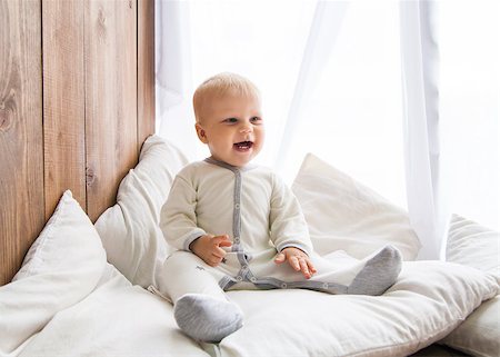 Toddler smiling cheerfully and looking away sitting on white feather with pillows near wooden wall Fotografie stock - Microstock e Abbonamento, Codice: 400-08790176