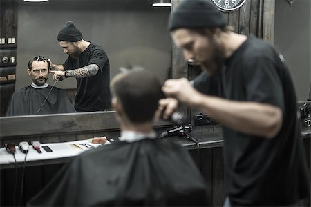 Matchless barber with a beard and a tattoo is cutting the hair of his bearded client in the barbershop. He is using a cutting comb and a hair clipper. They both reflected in a mirror. Horizontal. Stock Photo - Budget Royalty-Free & Subscription, Code: 400-08809917