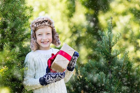 snow cosy - beautiful smiling boy in trapper hat, sweater and mittens holding nicely wrapped christmas gift by the tree or in the forest enjoying snowy cold winter weather, holiday or happiness concept Stock Photo - Budget Royalty-Free & Subscription, Code: 400-08815275