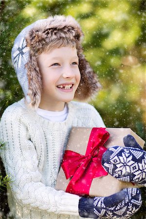 snow cosy - beautiful smiling boy in trapper hat, sweater and mittens holding nicely wrapped christmas gift by the tree or in the forest enjoying snowy cold winter weather, holiday or happiness concept Stock Photo - Budget Royalty-Free & Subscription, Code: 400-08815278