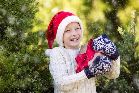 snow cosy - beautiful smiling boy in santa's hat, sweater and mittens holding nicely wrapped christmas gift by the tree or in the forest enjoying snowy cold winter weather, holiday or happiness concept Stock Photo - Budget Royalty-Free & Subscription, Code: 400-08815279