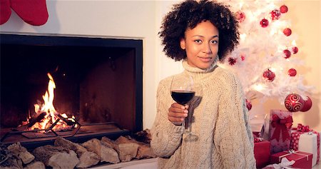 Pretty african american woman with huge afro haircut holds glass with red wine. She sitting next to fireplace and white christmas tree. She wearing winter woolen beige sweater. Foto de stock - Super Valor sin royalties y Suscripción, Código: 400-08811128