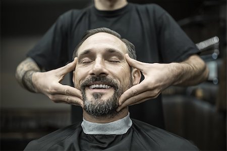 Smiling man with a beard and closed eyes in a black cutting hair cape in the barbershop. Barber with a tattoo in a black T-shirt is doing him a face massage. Closeup. Horizontal. Stock Photo - Budget Royalty-Free & Subscription, Code: 400-08813806