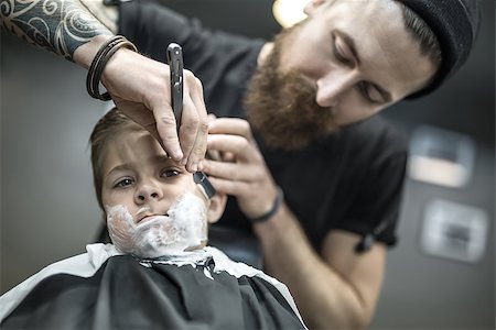 simsearch:673-02140507,k - Small kid with a shaving foam on the face in the barbershop. He wears a black salon cape. Bearded barber with a tattoo is shaving boy's face with the help of the straight razor. Low aperture photo. Foto de stock - Royalty-Free Super Valor e Assinatura, Número: 400-08861656