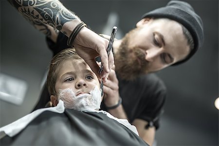 simsearch:673-02140507,k - Little kid with a shaving foam on the face in the barbershop. He wears a black salon cape. Bearded barber with a tattoo is shaving boy's face with the help of the straight razor. Low aperture photo. Foto de stock - Royalty-Free Super Valor e Assinatura, Número: 400-08861650