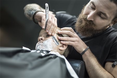 simsearch:673-02140507,k - Cute kid with a shaving foam on the face in the barbershop. He wears a black salon cape. Bearded barber with a tattoo is shaving boy's face with the help of the straight razor. Low aperture photo. Foto de stock - Royalty-Free Super Valor e Assinatura, Número: 400-08861651