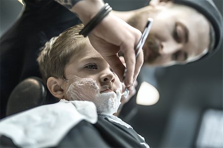 simsearch:673-02140507,k - Brutal kid with a shaving foam on the face in the barbershop. He wears a black salon cape. Bearded barber with a tattoo is shaving boy's face with the help of the straight razor. Low aperture photo. Foto de stock - Royalty-Free Super Valor e Assinatura, Número: 400-08861653