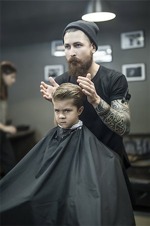 simsearch:673-02140507,k - Saucy little boy in a black salon cape in the barbershop. Bearded barber with a tattoo in a black cap does kid the hairstyle. Vertical. Foto de stock - Royalty-Free Super Valor e Assinatura, Número: 400-08862475