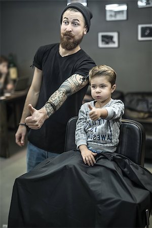 simsearch:673-02140507,k - Happy small kid with the new hairstyle and the bearded tattooed barber are doing grimaces and showing the thumbs up sign. Boy wears a gray sweatshirt. Hairdresser wears a black T-shirt and cap, jeans. Foto de stock - Royalty-Free Super Valor e Assinatura, Número: 400-08862477