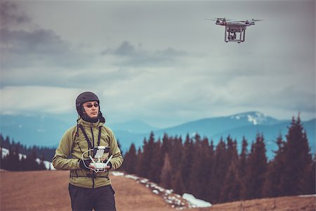 Young man in green jacket operating a drone using a remote controller. Ski resort in the background, winter landscape with pine tree forest and mountains. Vintage toned image. Bukovel, Carpathians, Ukraine, Europe. Exploring beauty world Stock Photo - Budget Royalty-Free & Subscription, Code: 400-08863887