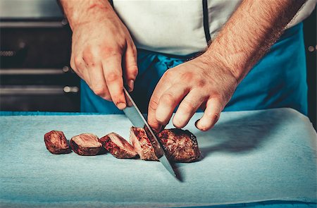 simsearch:614-03903584,k - Chef in hotel or restaurant kitchen dressed in blue apron cooking, only hands, he is cutting meat or steak with big sharp knife Foto de stock - Royalty-Free Super Valor e Assinatura, Número: 400-08863893