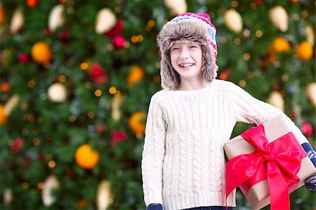 snow cosy - positive smiling child in trapper hat and warm winter clothes holding huge christmas gift enjoying christmas time by the tree Stock Photo - Budget Royalty-Free & Subscription, Code: 400-08887763