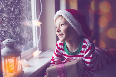 snow cosy - happy smiling boy holding nicely wrapped gift sitting cozy at home looking out the window while snowing waiting for christmas miracle, christmas lights and decorations, holiday concept, toned image Stock Photo - Budget Royalty-Free & Subscription, Code: 400-08888144