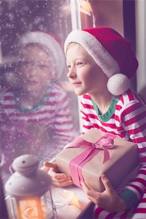 snow cosy - happy smiling boy holding nicely wrapped gift sitting cozy at home looking out the window while snowing waiting for christmas miracle, christmas lights and decorations, holiday concept, toned image Stock Photo - Budget Royalty-Free & Subscription, Code: 400-08888143