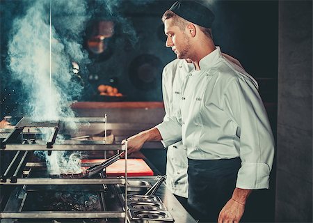 Young white chef in black apron and hat standing near the brazier whith coals. Man cooking beef steak in the interior of modern professional kitchen Stock Photo - Budget Royalty-Free & Subscription, Code: 400-08893311