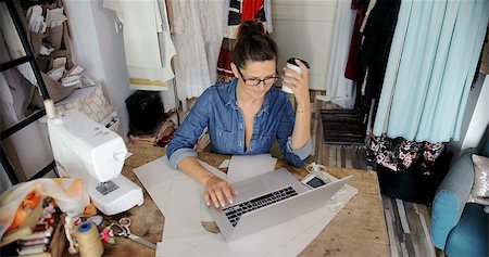 simsearch:400-04339323,k - Young woman in glasses sitting at table with coffee and using laptop while working in tailor's shop. Foto de stock - Royalty-Free Super Valor e Assinatura, Número: 400-08963157