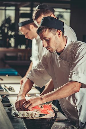 simsearch:400-08192842,k - Three young chefs dressed in white uniform decorate ready dish in restaurant. They are working on maki rolls. Preparing traditional japanese sushi set in interior of modern professional kitchen Foto de stock - Super Valor sin royalties y Suscripción, Código: 400-09028929