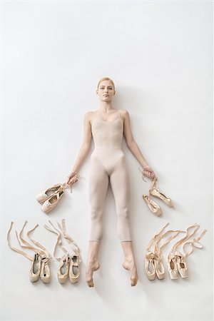 simsearch:400-05739749,k - Attractive ballerina lies with stretched legs on the white floor and holds pointe shoes in the studio. She wears a light dance wear. On the sides of her legs there are ballet shoes. Top view photo. Photographie de stock - Aubaine LD & Abonnement, Code: 400-09029135