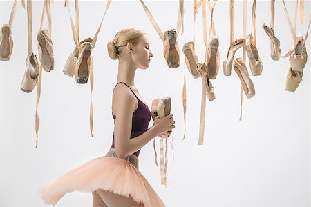 simsearch:400-07660010,k - Sensual ballerina in a violet top and a peach tutu stands sideways on the light background in the studio. Around her there are many hanging beige pointe shoes. She holds one pair in the hands. Stock Photo - Budget Royalty-Free & Subscription, Code: 400-09029323