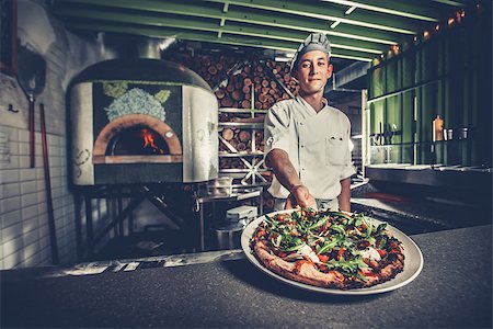 simsearch:6126-08635695,k - Food concept. Preparing traditional italian pizza. Young smiling chef in white uniform and gray hat show ready dish with green rucola herbs in interior of modern restaurant kitchen. Ready to eat. Foto de stock - Super Valor sin royalties y Suscripción, Código: 400-09064059