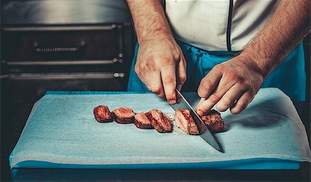 simsearch:614-03903584,k - Food concept. Preparing traditional beef steak in interior of modern restaurant kitchen. Chef dressed in blue apron cooking, only hands, he is cutting meat steak with big sharp knife on cutting board Foto de stock - Royalty-Free Super Valor e Assinatura, Número: 400-09062787